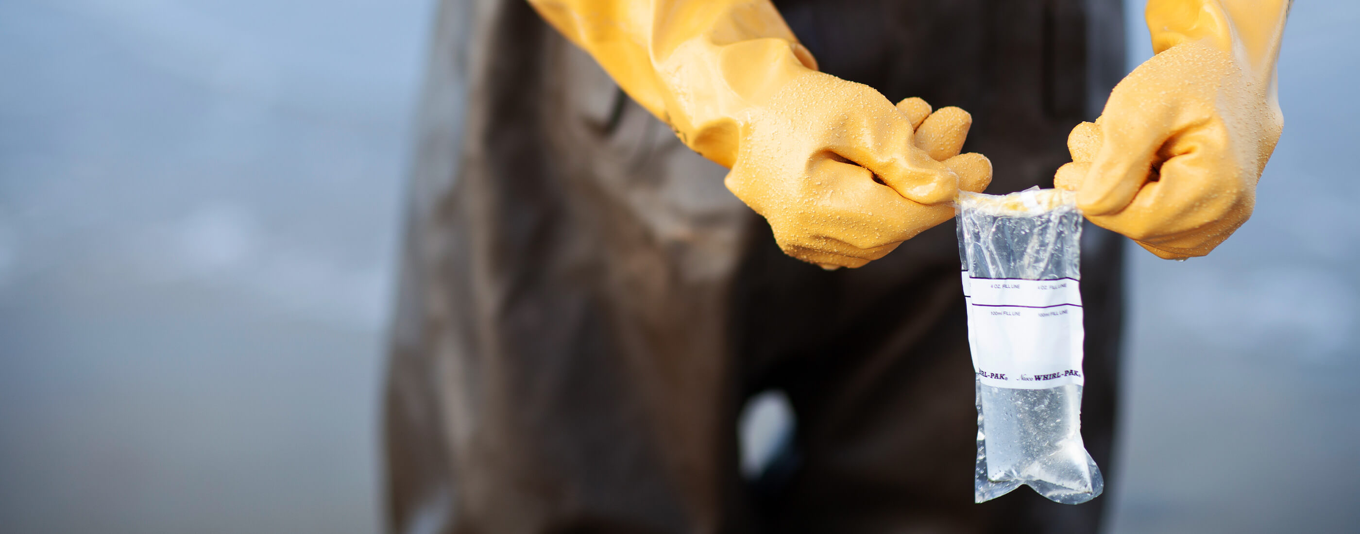 Hands in yellow safety gloves hold up a filled water sample along the beach.